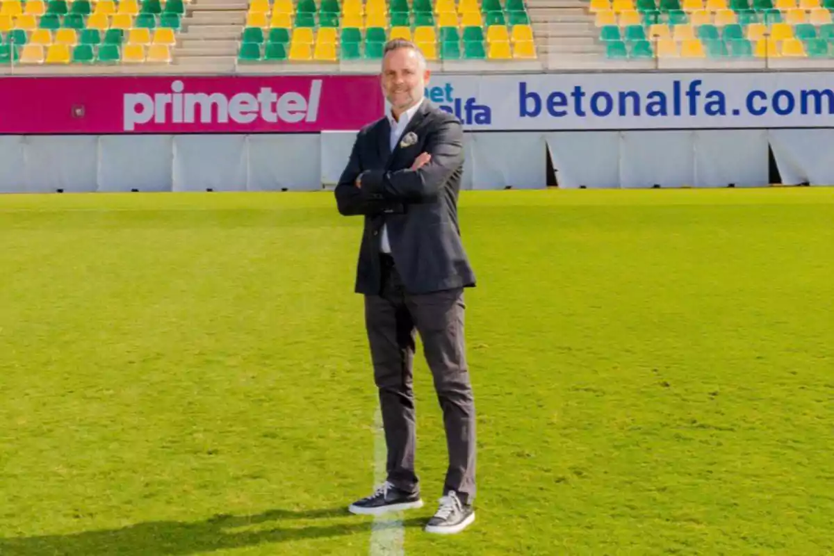 Hombre de pie con los brazos cruzados en el centro de un campo de fútbol con gradas de asientos amarillos y verdes al fondo Hombre de pie con los brazos cruzados en el centro de un campo de fútbol con gradas de asientos amarillos y verdes al fondo