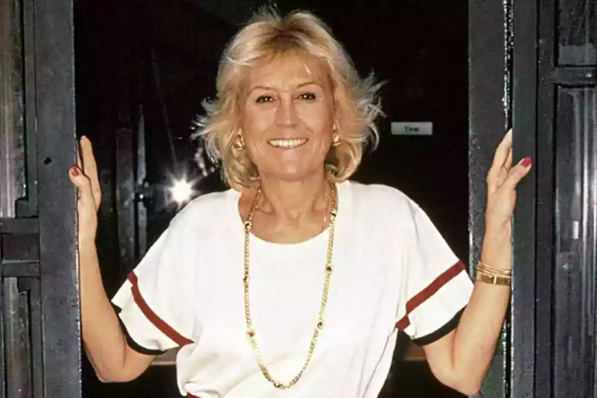 Mujer rubia de mediana edad sonriente con camiseta blanca y collar dorado de pie en una puerta Mujer rubia de mediana edad sonriente con camiseta blanca y collar dorado de pie en una puerta