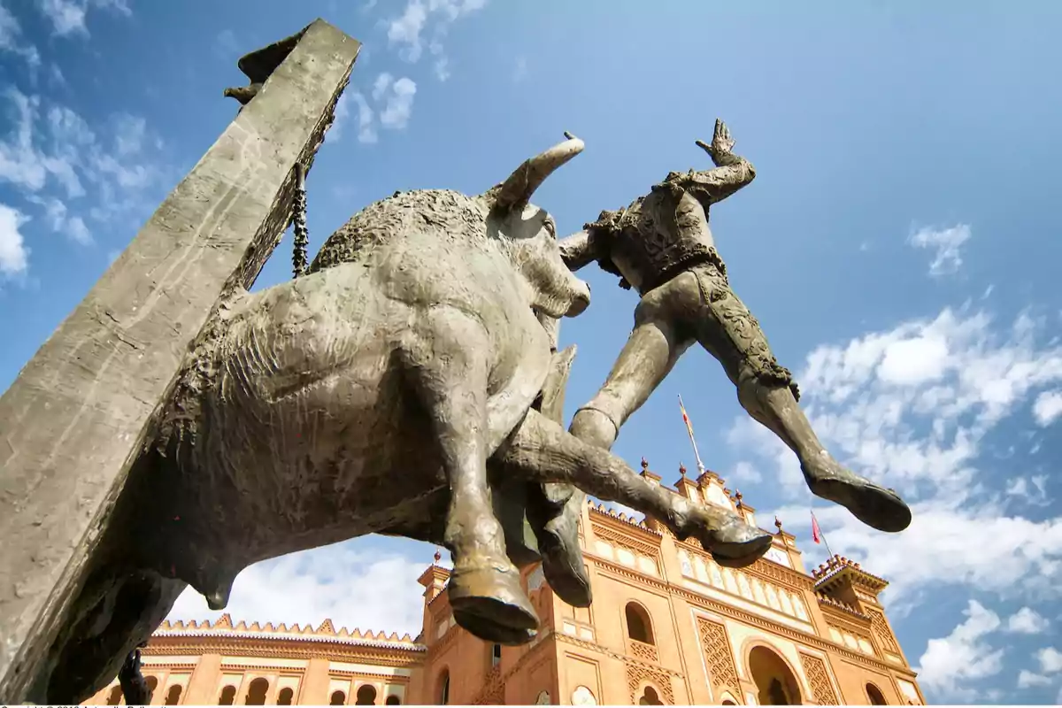 Escultura de un toro embistiendo a un torero frente a la plaza de toros de Las Ventas en Madrid bajo un cielo azul con algunas nubes Escultura de un toro embistiendo a un torero frente a la plaza de toros de Las Ventas en Madrid bajo un cielo azul con algunas nubes