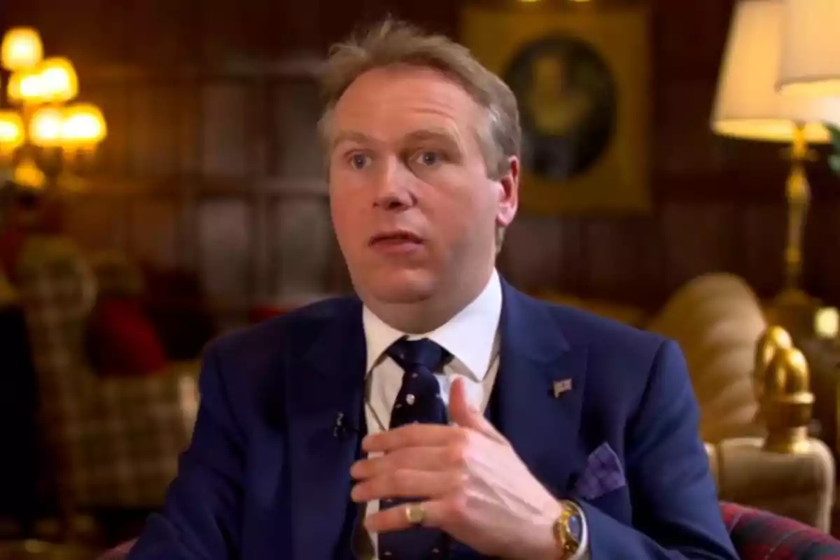 Grant Harrold de traje azul hablando en una sala elegantemente decorada con lámparas y muebles clásicos Grant Harrold de traje azul hablando en una sala elegantemente decorada con lámparas y muebles clásicos