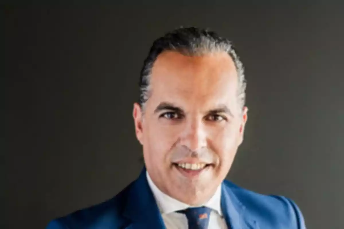 Hombre de mediana edad con cabello oscuro peinado hacia atrás vistiendo un traje azul y corbata frente a un fondo oscuro Hombre de mediana edad con cabello oscuro peinado hacia atrás vistiendo un traje azul y corbata frente a un fondo oscuro