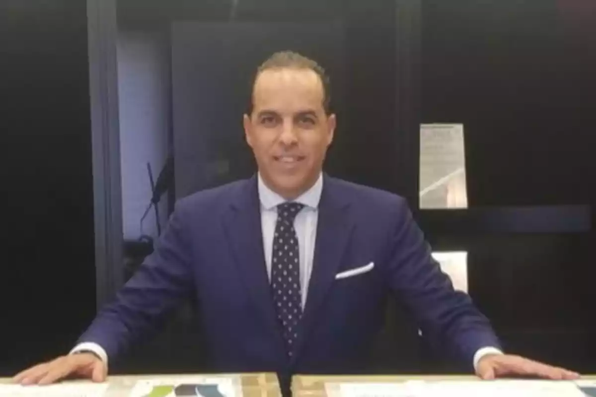 Hombre de traje azul y corbata de puntos posando frente a una mesa en un entorno de oficina Hombre de traje azul y corbata de puntos posando frente a una mesa en un entorno de oficina