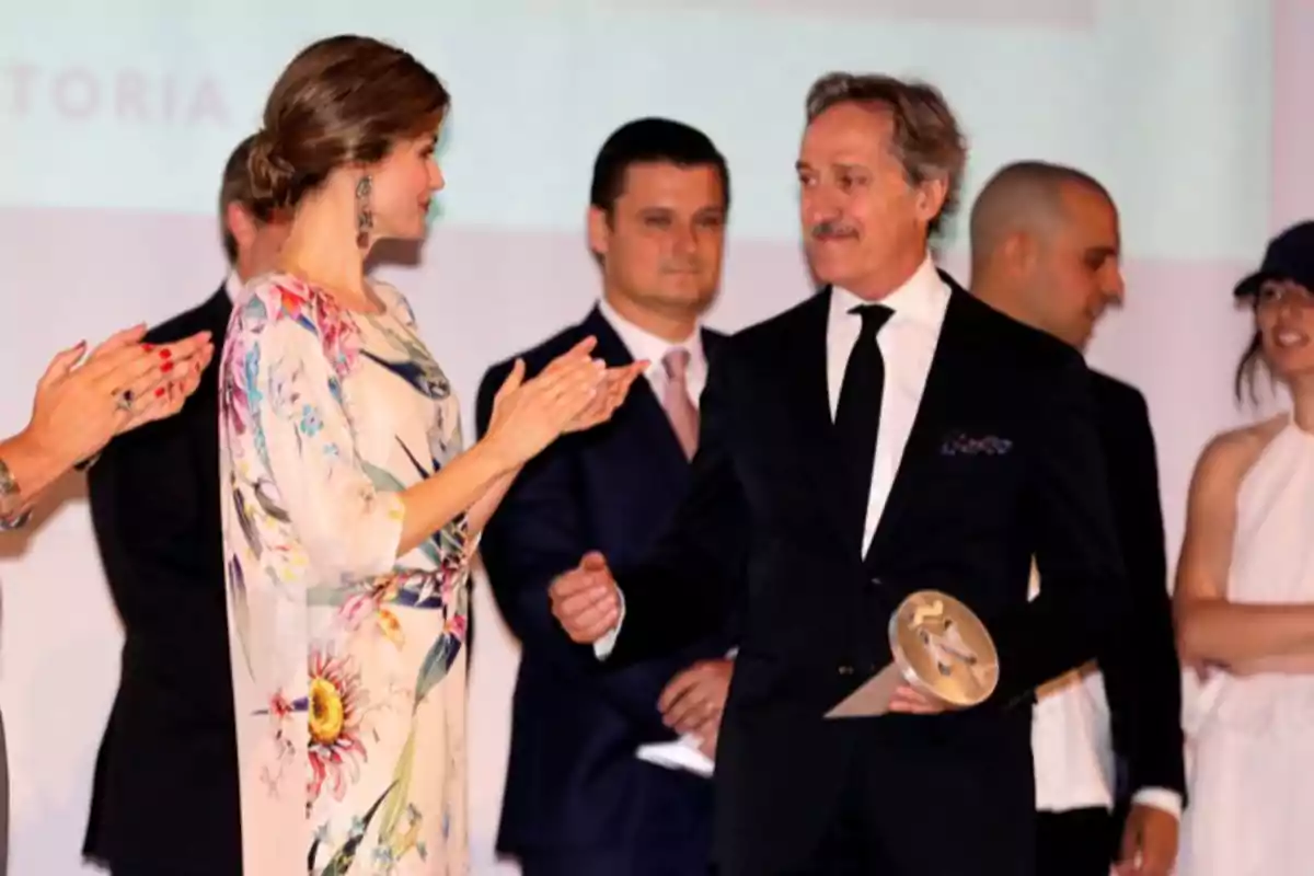 Un hombre con traje sostiene un premio mientras una mujer con vestido floral lo aplaude en un escenario rodeados de otras personas elegantemente vestidas Un hombre con traje sostiene un premio mientras una mujer con vestido floral lo aplaude en un escenario rodeados de otras personas elegantemente vestidas