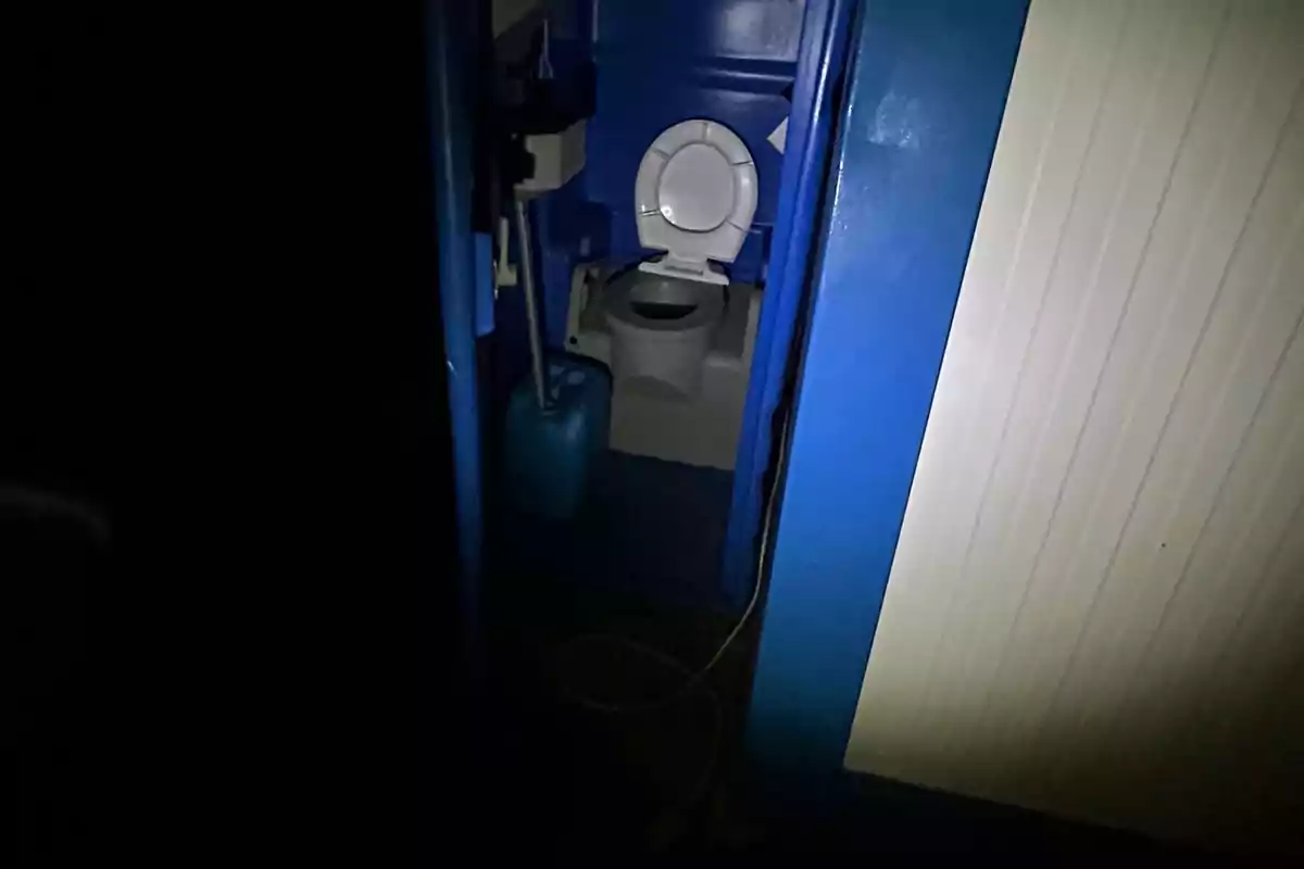 Baño portátil azul iluminado parcialmente en la oscuridad con la puerta entreabierta Baño portátil azul iluminado parcialmente en la oscuridad con la puerta entreabierta