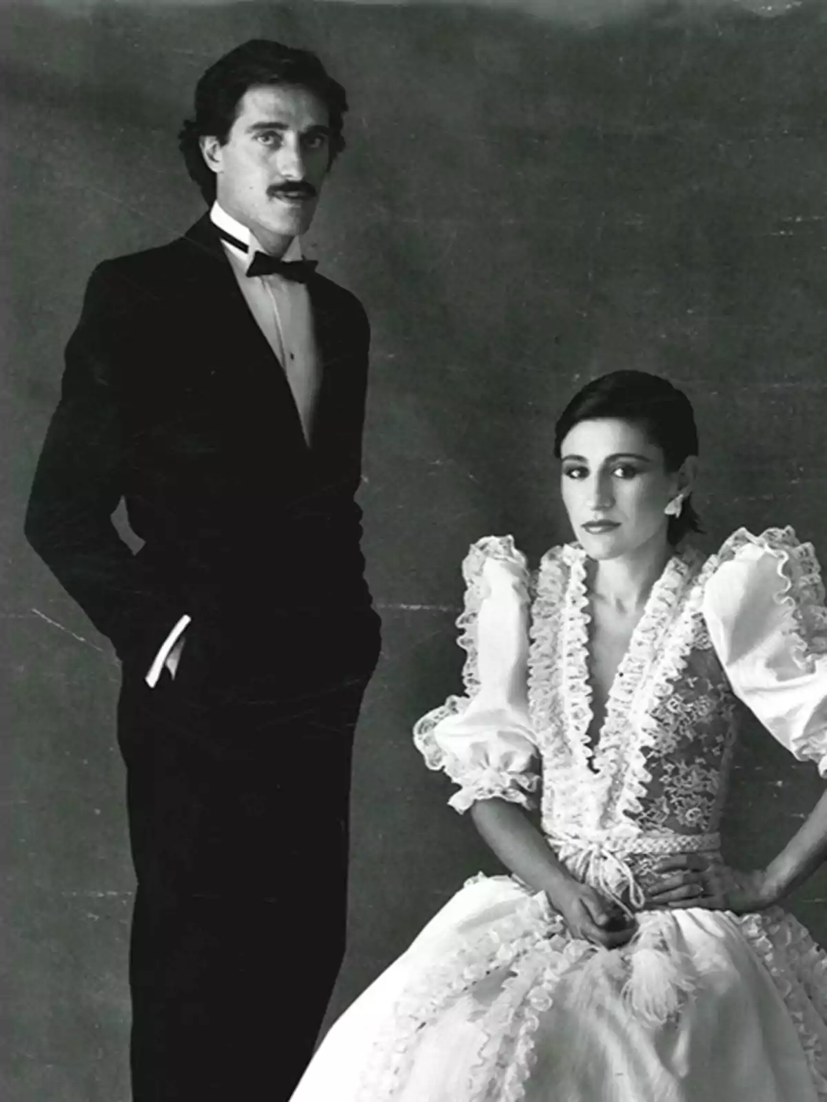 Pareja posando elegantemente en una fotografía en blanco y negro, el hombre lleva esmoquin y la mujer un vestido de novia con encaje y mangas abullonadas Pareja posando elegantemente en una fotografía en blanco y negro, el hombre lleva esmoquin y la mujer un vestido de novia con encaje y mangas abullonadas