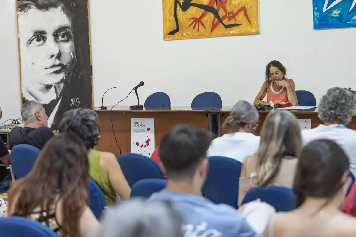 Una mujer lee en voz alta sentada en una mesa frente a un público en una sala decorada con pinturas y carteles de un festival literario. Una mujer lee en voz alta sentada en una mesa frente a un público en una sala decorada con pinturas y carteles de un festival literario.