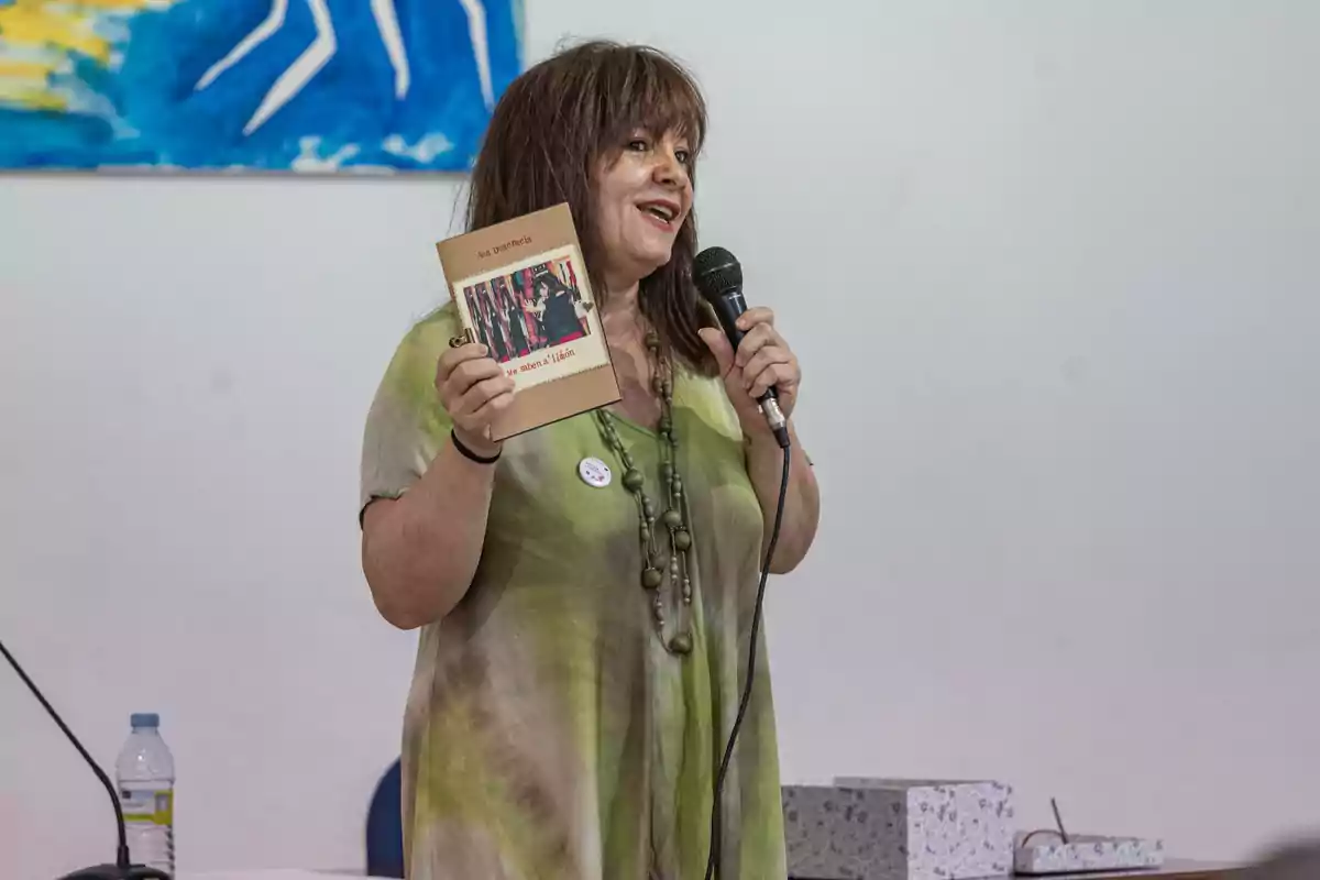 Mujer de cabello castaño sostiene un libro y un micrófono mientras habla en un evento en interiores, lleva un vestido verde y un collar largo, al fondo hay una pintura azul y una mesa con una botella de agua y una caja.