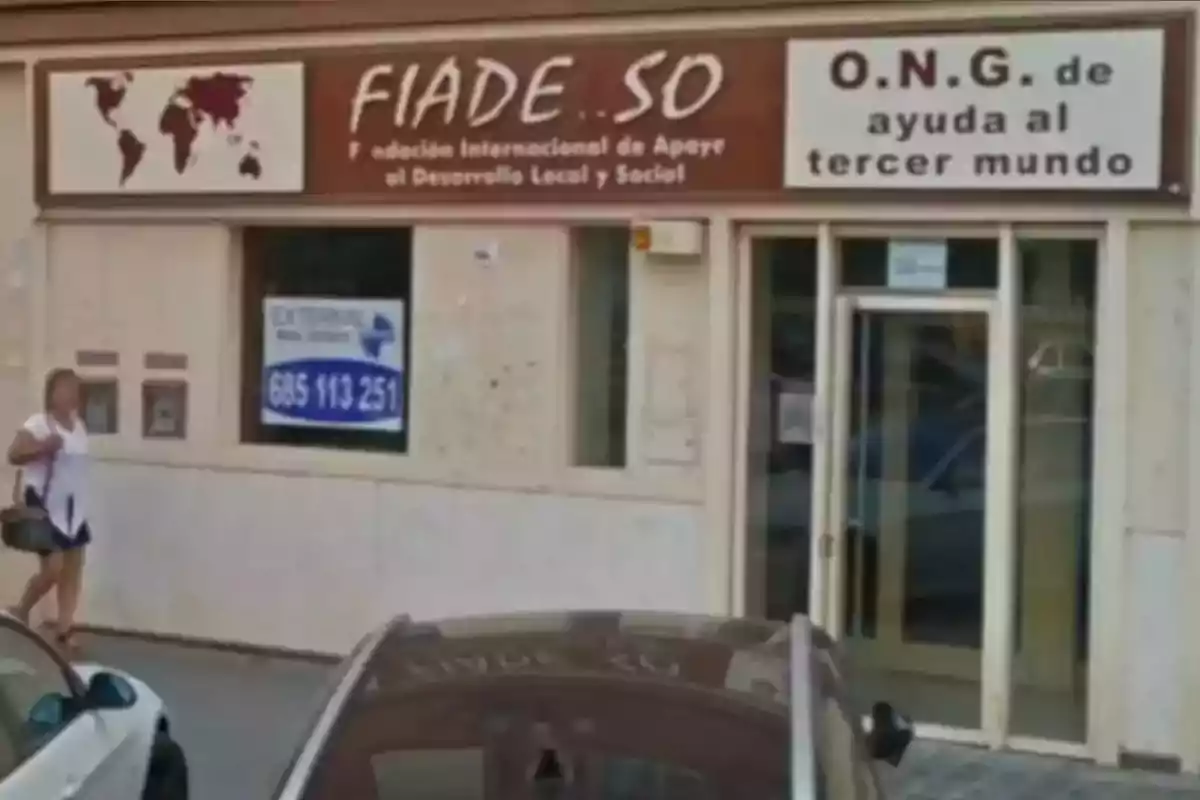 Fachada de una organización no gubernamental con letreros que indican ayuda al desarrollo local y social, una mujer caminando por la acera y autos estacionados frente al edificio Fachada de una organización no gubernamental con letreros que indican ayuda al desarrollo local y social, una mujer caminando por la acera y autos estacionados frente al edificio