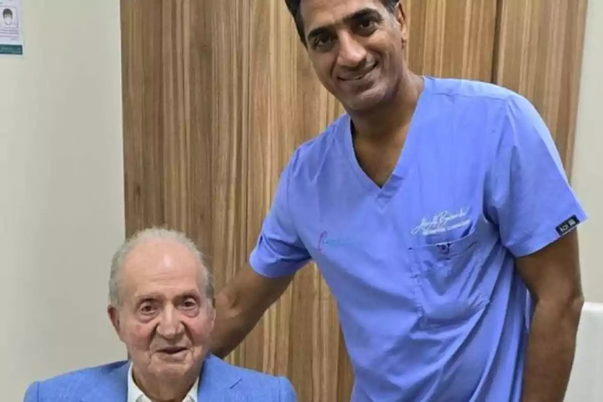Un hombre mayor con saco azul está sentado junto a un hombre con uniforme médico azul que sonríe y lo acompaña en una consulta médica Un hombre mayor con saco azul está sentado junto a un hombre con uniforme médico azul que sonríe y lo acompaña en una consulta médica