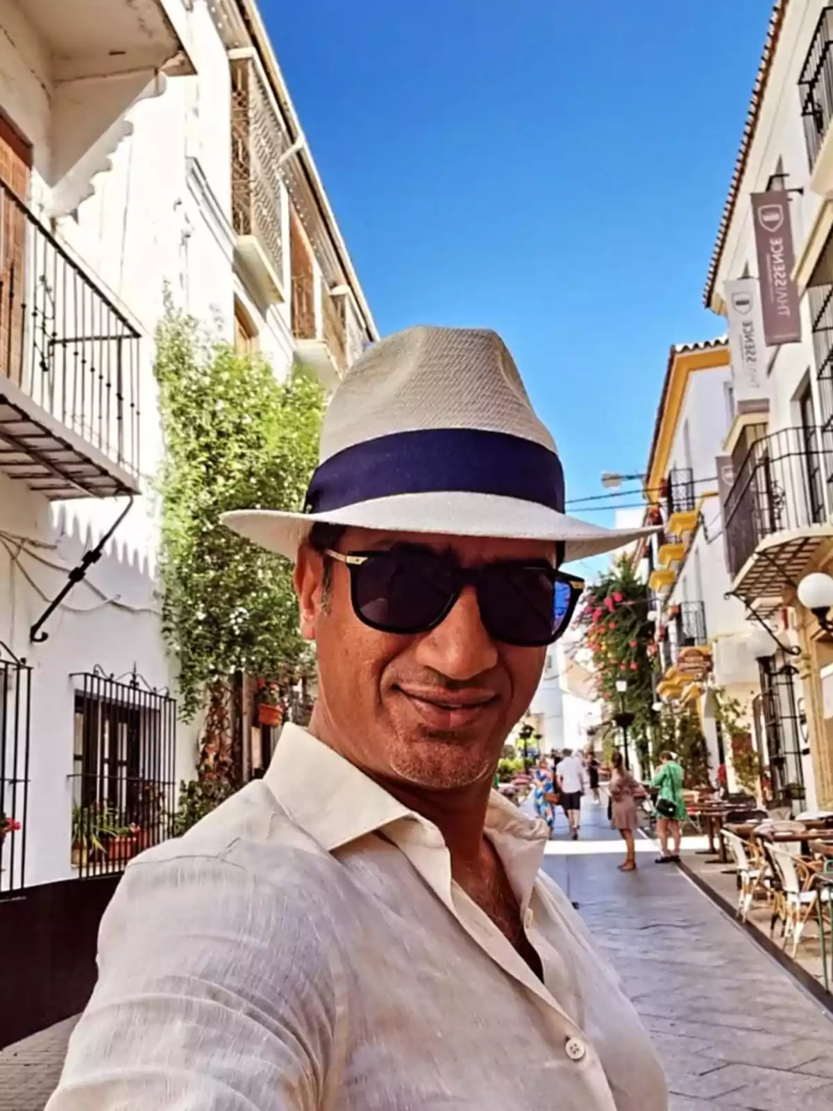 Hombre con sombrero y gafas de sol tomándose una selfie en una calle peatonal con edificios blancos y cielo despejado Hombre con sombrero y gafas de sol tomándose una selfie en una calle peatonal con edificios blancos y cielo despejado
