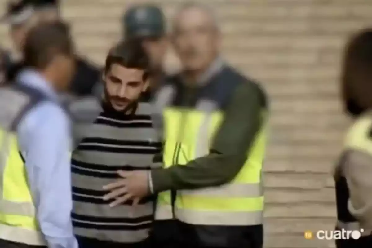 Un hombre con suéter de rayas es escoltado por agentes con chalecos reflectantes en una zona exterior Un hombre con suéter de rayas es escoltado por agentes con chalecos reflectantes en una zona exterior