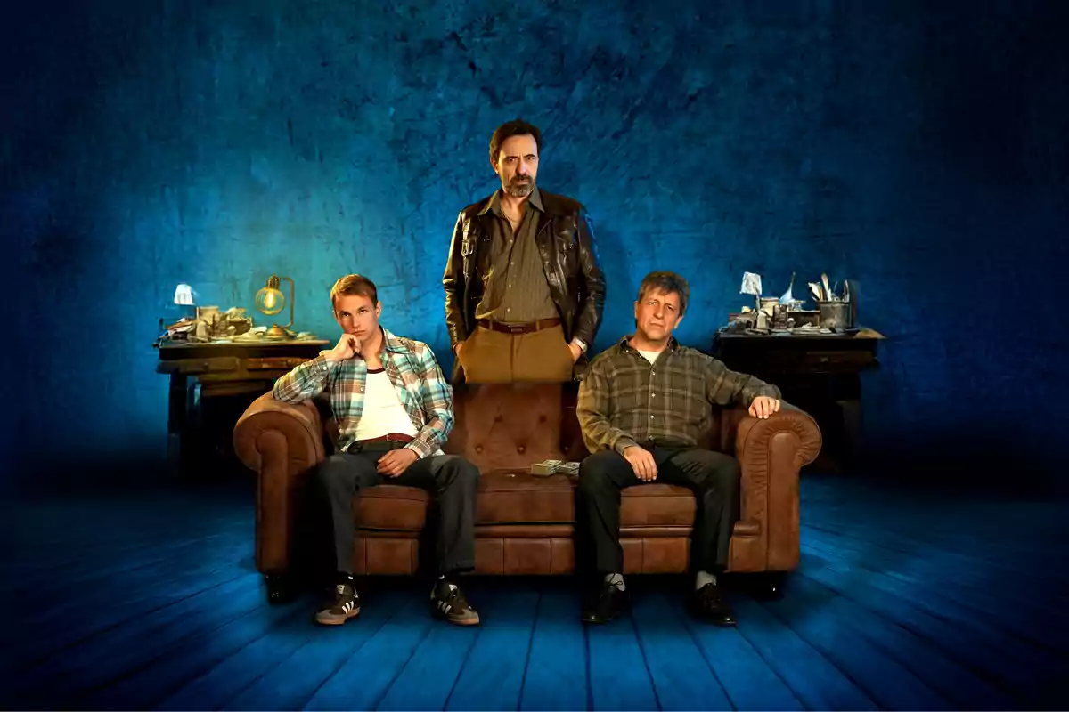 Tres hombres posan en un ambiente teatral con fondo azul, dos de ellos están sentados en un sofá marrón y el tercero está de pie detrás de ellos, al fondo hay escritorios con lámparas y objetos desordenados.