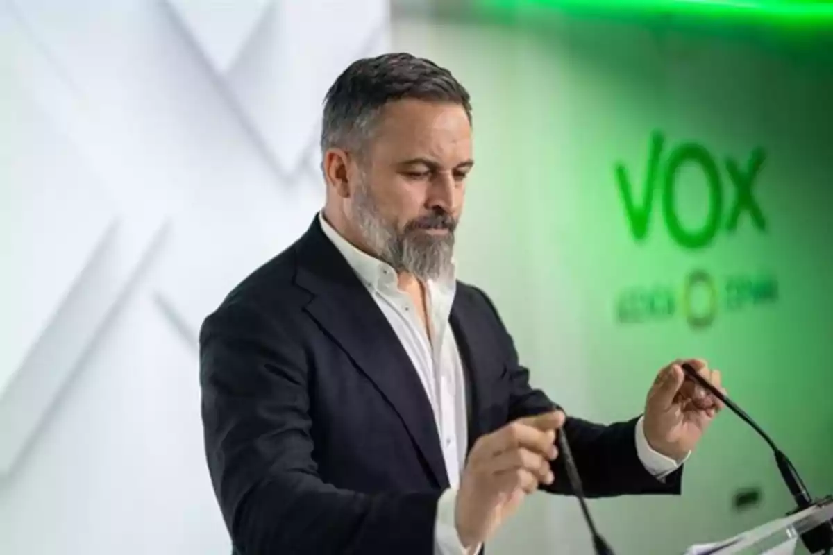 Hombre de barba y cabello corto con traje oscuro y camisa blanca de pie frente a un atril con micrófonos y el logo de VOX en el fondo Hombre de barba y cabello corto con traje oscuro y camisa blanca de pie frente a un atril con micrófonos y el logo de VOX en el fondo