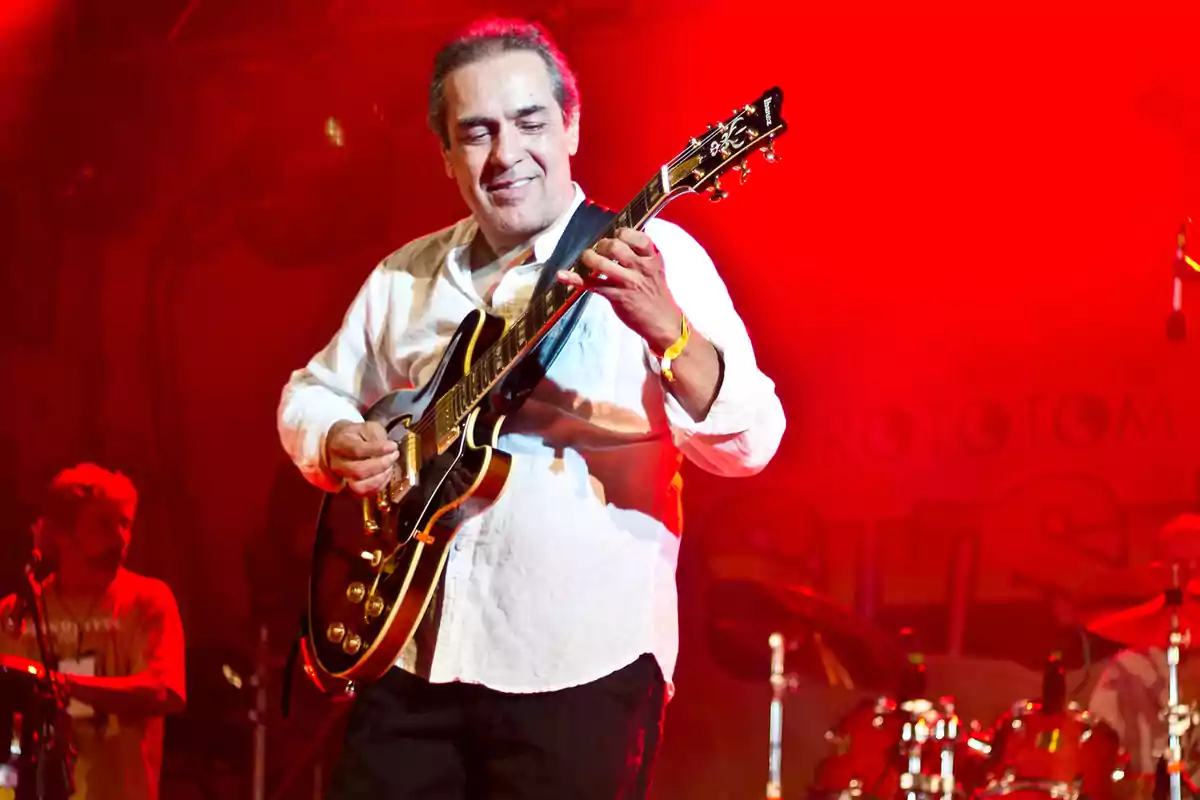Hombre tocando una guitarra eléctrica en el escenario con luces rojas de fondo y expresión sonriente Hombre tocando una guitarra eléctrica en el escenario con luces rojas de fondo y expresión sonriente