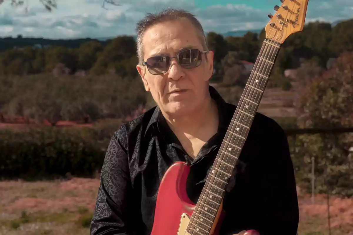 Hombre de mediana edad con gafas de sol y camisa oscura sosteniendo una guitarra eléctrica roja en un entorno natural
