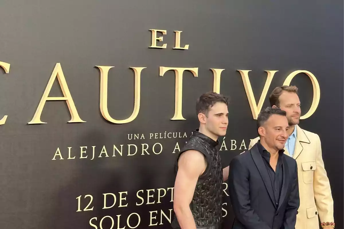 Tres hombres posan juntos en una alfombra roja frente a un fondo promocional de la película El Cautivo Tres hombres posan juntos en una alfombra roja frente a un fondo promocional de la película El Cautivo
