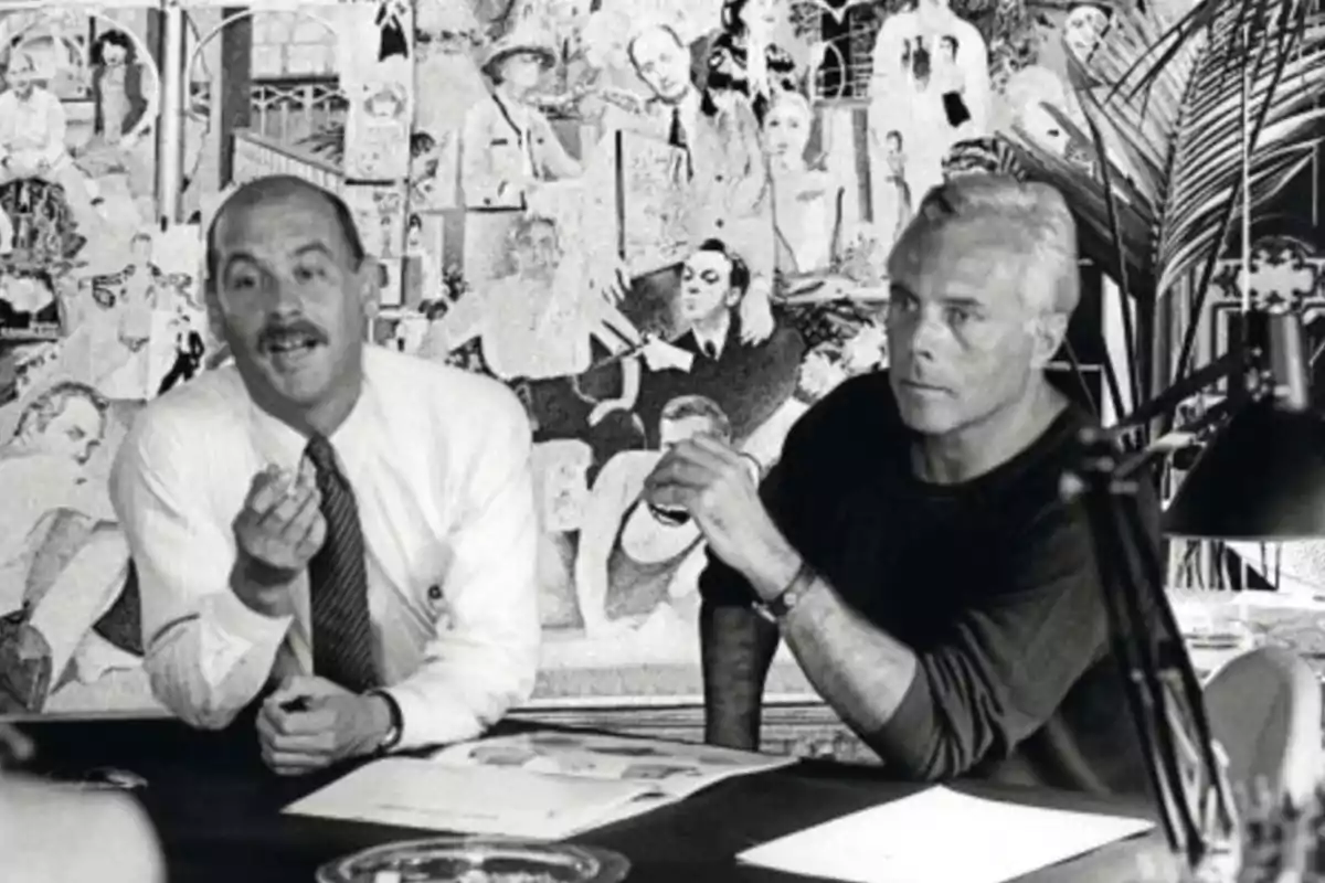 Dos hombres sentados en una mesa de trabajo con papeles y una lámpara, al fondo un mural con ilustraciones y retratos en blanco y negro Dos hombres sentados en una mesa de trabajo con papeles y una lámpara, al fondo un mural con ilustraciones y retratos en blanco y negro