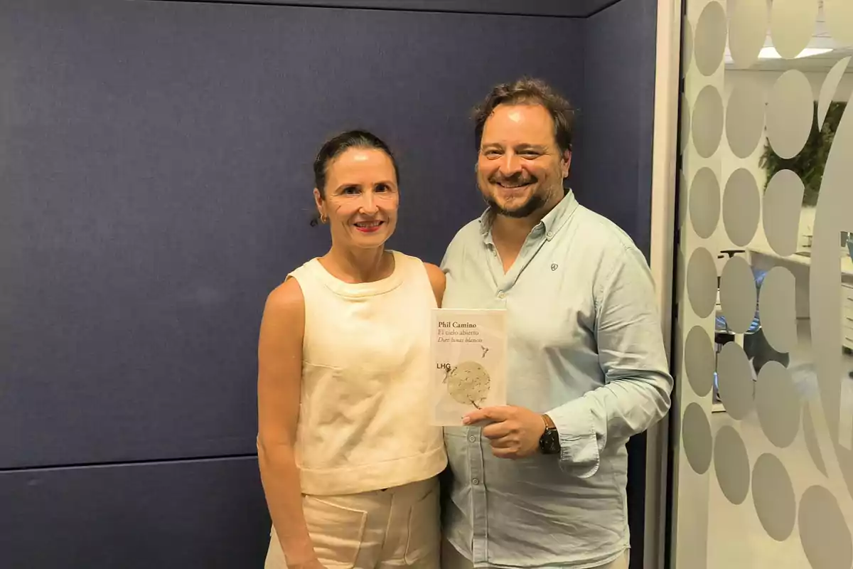Dos personas sonríen mientras posan juntas en una oficina moderna y una de ellas sostiene un libro Dos personas sonríen mientras posan juntas en una oficina moderna y una de ellas sostiene un libro