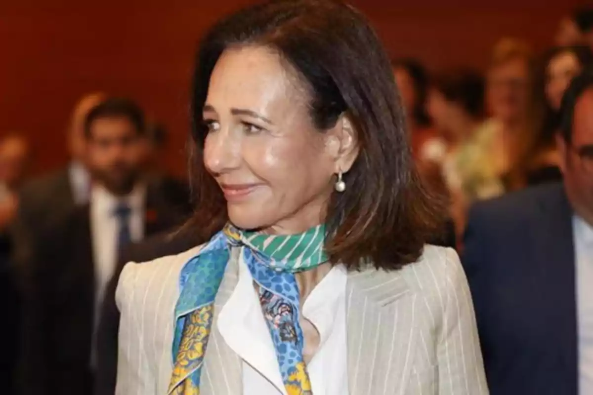 Mujer de cabello castaño sonriente con un pañuelo colorido en el cuello y saco claro en un evento formal con personas al fondo desenfocadas Mujer de cabello castaño sonriente con un pañuelo colorido en el cuello y saco claro en un evento formal con personas al fondo desenfocadas