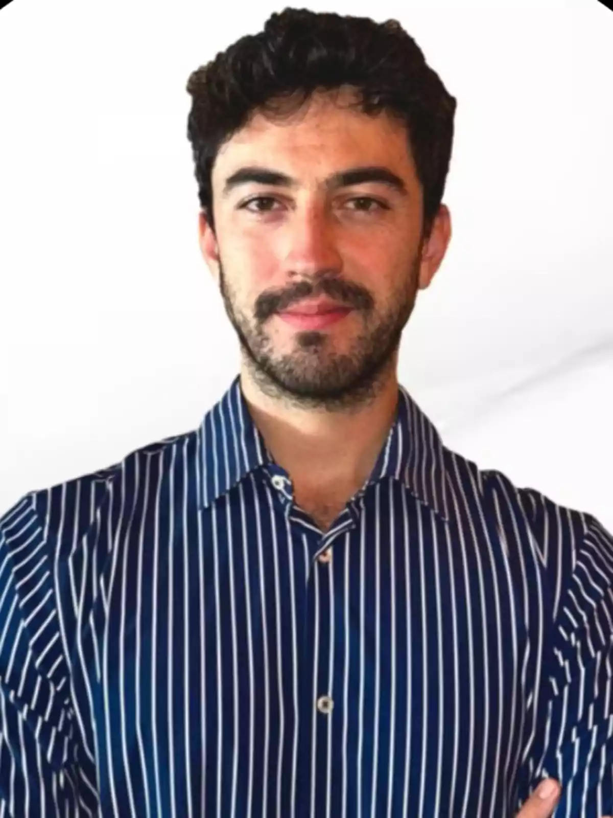 Hombre joven de cabello oscuro y barba corta vistiendo una camisa azul de rayas blancas posando frente a un fondo blanco Hombre joven de cabello oscuro y barba corta vistiendo una camisa azul de rayas blancas posando frente a un fondo blanco
