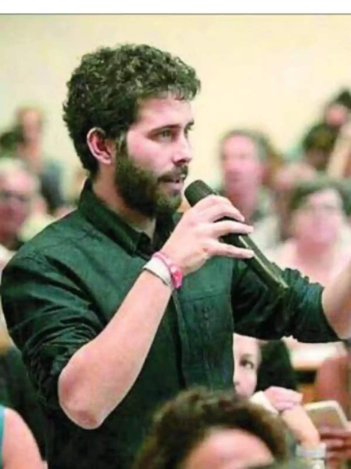 Un hombre joven con barba y cabello rizado sostiene un micrófono mientras habla en un evento con varias personas al fondo Un hombre joven con barba y cabello rizado sostiene un micrófono mientras habla en un evento con varias personas al fondo