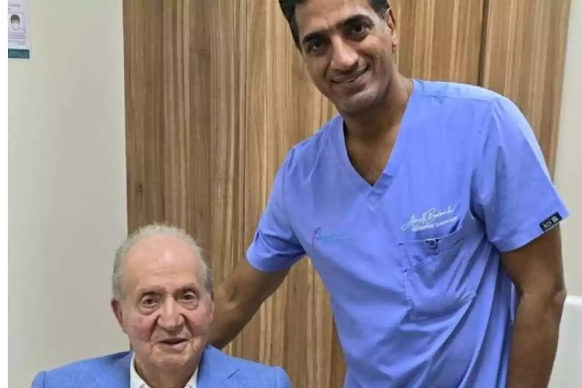 Dos hombres posan juntos en una habitación, uno de ellos está sentado y lleva un saco azul, mientras que el otro está de pie y viste un uniforme médico azul claro.