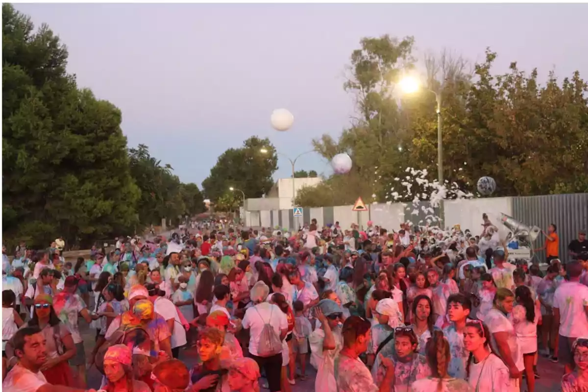 Multitud de personas celebrando una fiesta al aire libre con polvos de colores y espuma en una calle rodeada de árboles al atardecer Multitud de personas celebrando una fiesta al aire libre con polvos de colores y espuma en una calle rodeada de árboles al atardecer