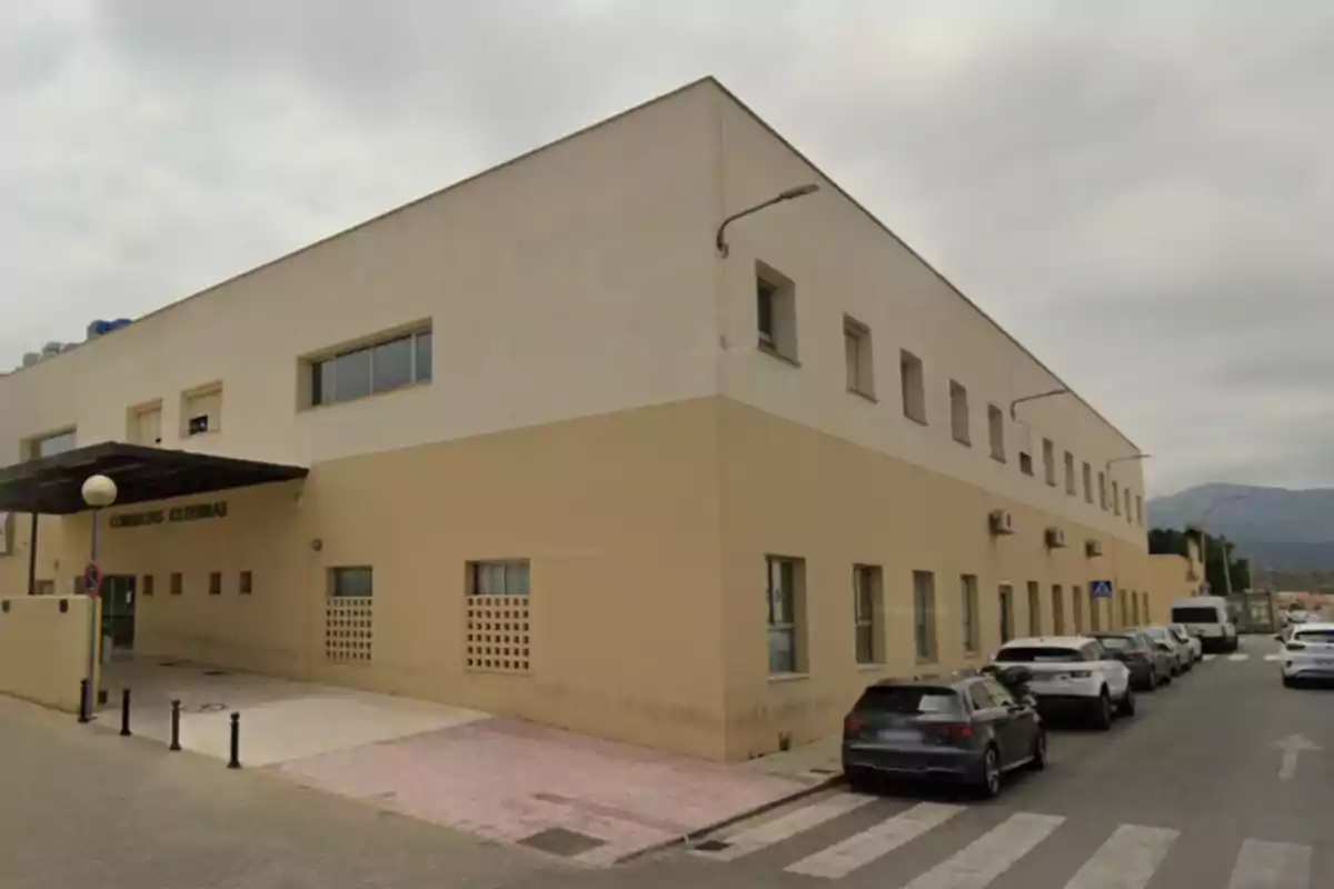 Edificio de dos plantas de color beige con letrero de consultas externas en la entrada principal y varios coches estacionados en la calle junto a una acera