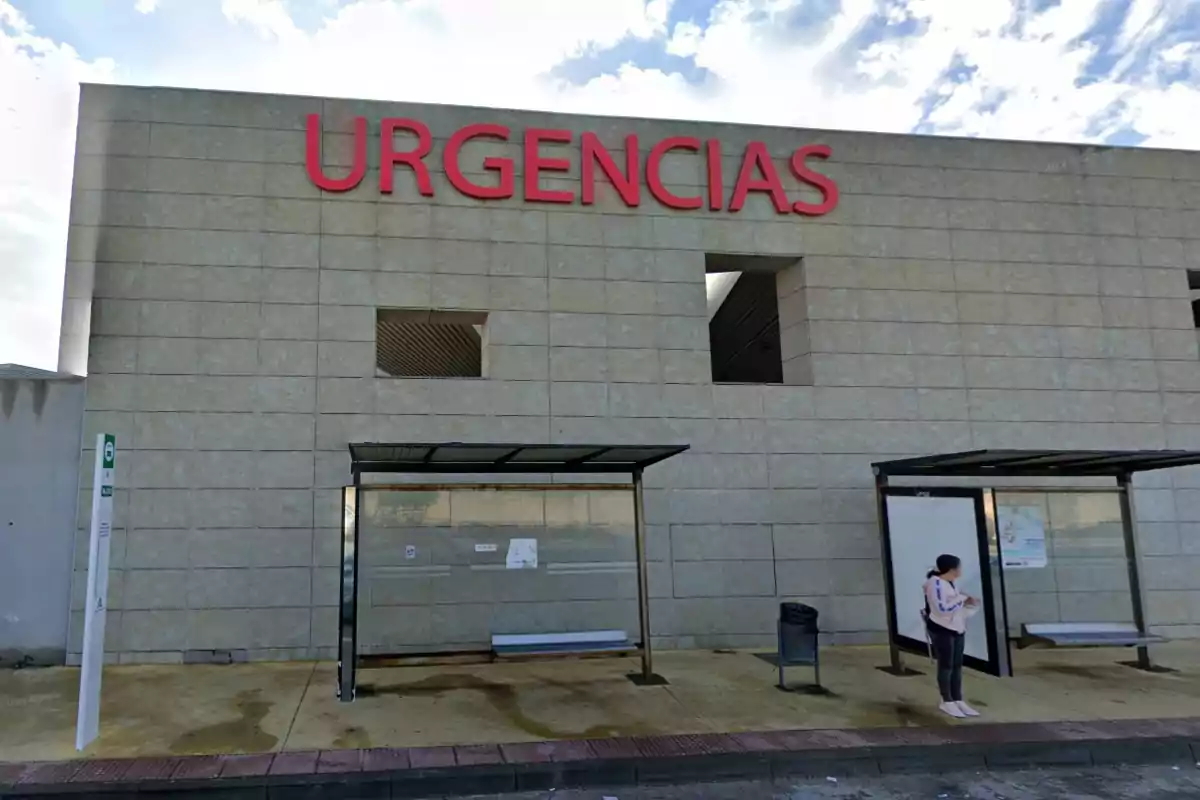 Fachada de un hospital con la palabra URGENCIAS en letras rojas y una persona esperando en una parada de autobús frente al edificio Fachada de un hospital con la palabra URGENCIAS en letras rojas y una persona esperando en una parada de autobús frente al edificio