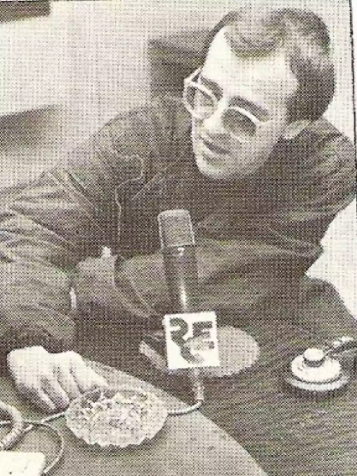 Hombre con gafas grandes hablando frente a un micrófono en una mesa de radio Hombre con gafas grandes hablando frente a un micrófono en una mesa de radio