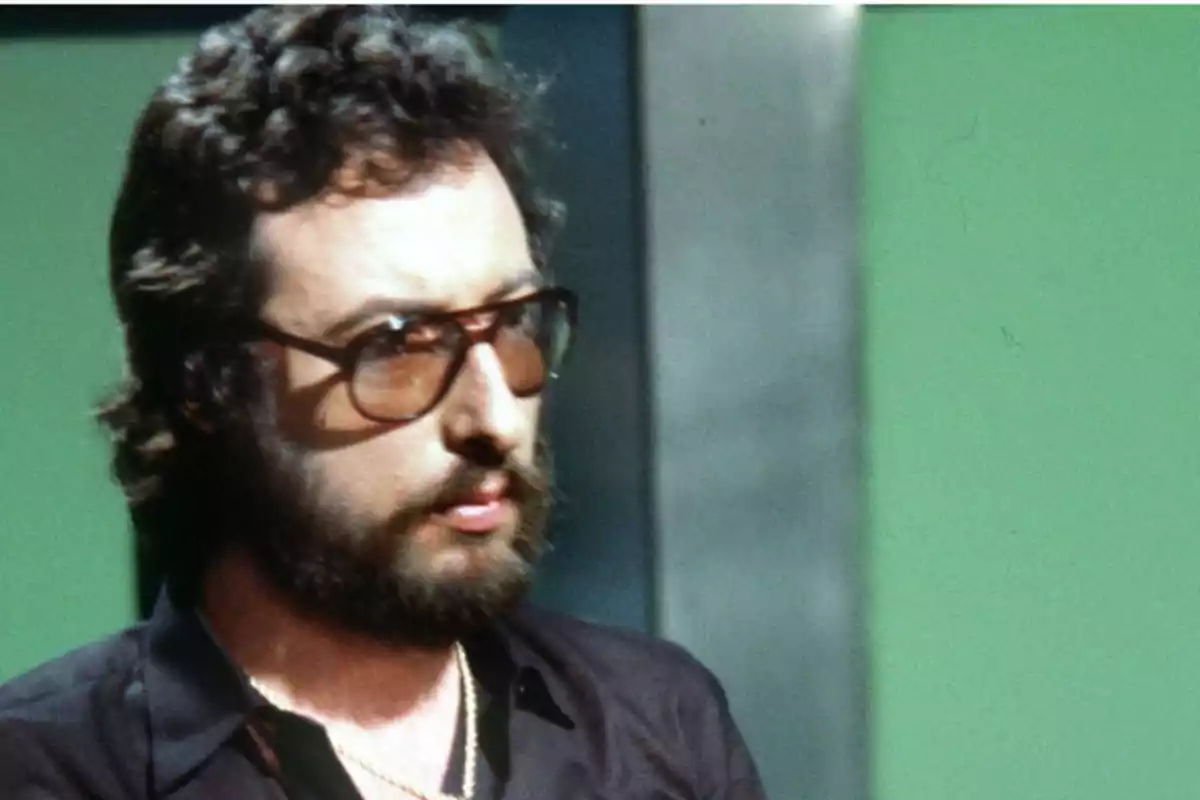 Hombre de cabello rizado y barba usando gafas grandes y camisa oscura frente a un fondo verde Hombre de cabello rizado y barba usando gafas grandes y camisa oscura frente a un fondo verde