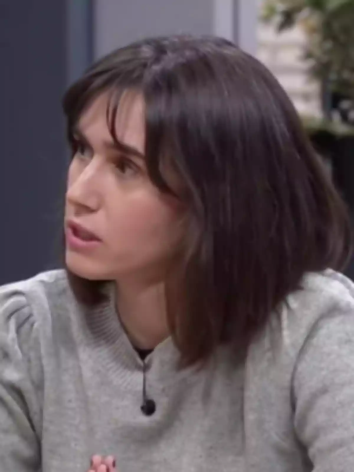 Mujer de cabello castaño y liso con suéter gris sentada en un ambiente interior mientras parece estar hablando Mujer de cabello castaño y liso con suéter gris sentada en un ambiente interior mientras parece estar hablando