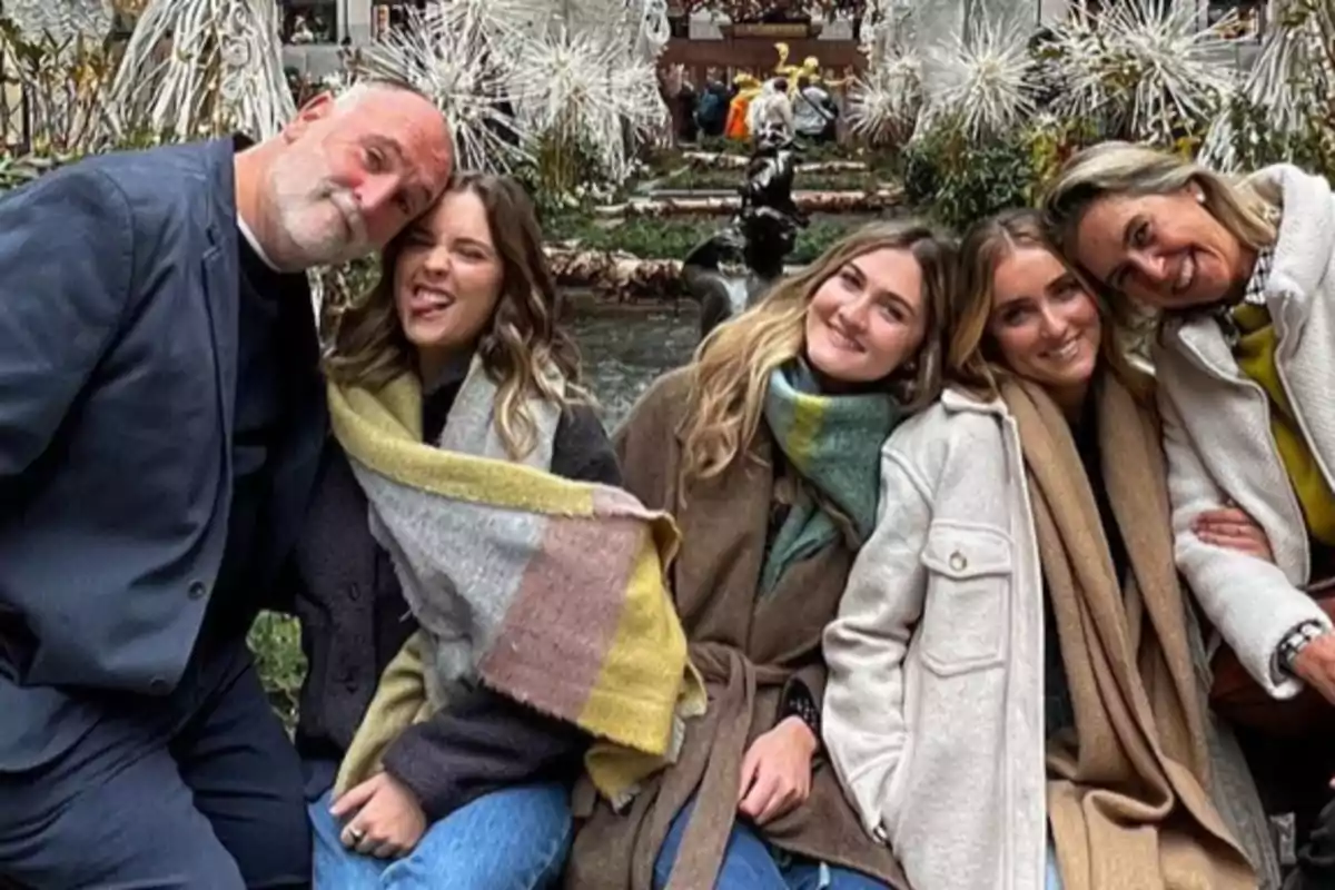 Cinco personas sentadas juntas al aire libre sonriendo y abrazándose con decoraciones invernales de fondo Cinco personas sentadas juntas al aire libre sonriendo y abrazándose con decoraciones invernales de fondo