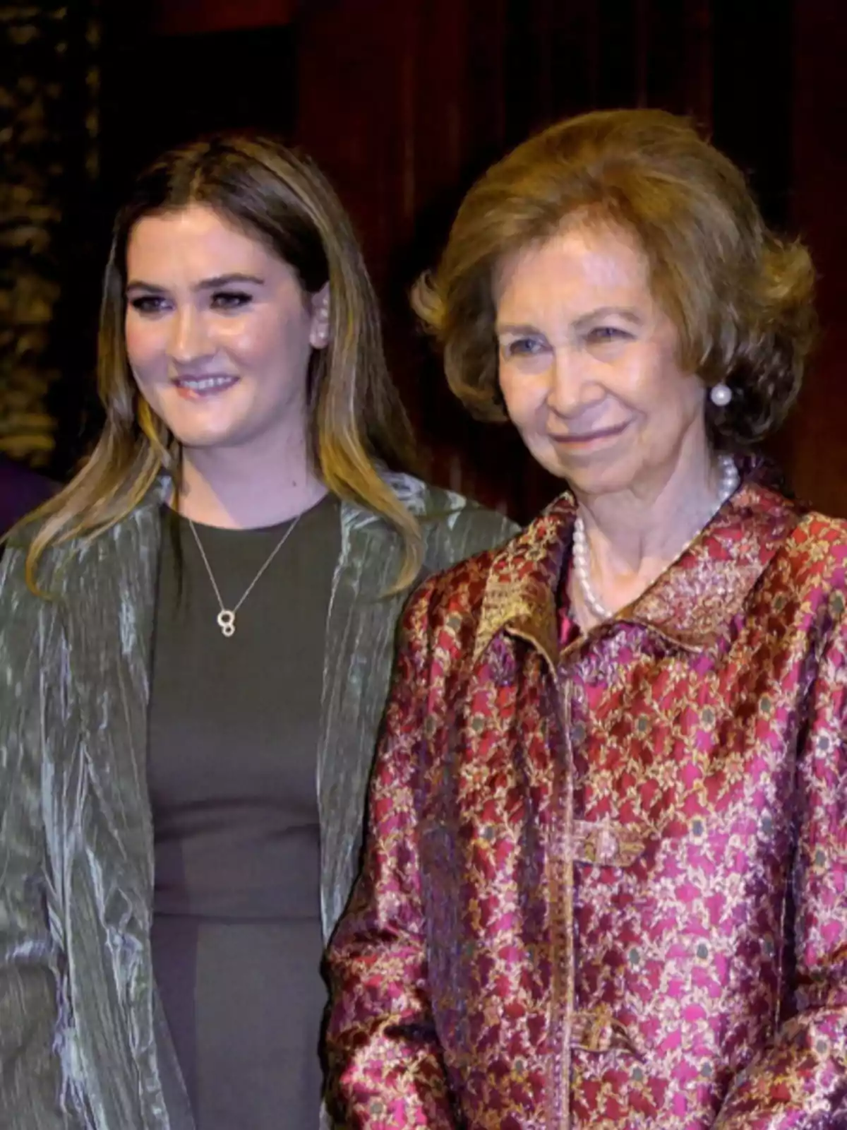 Dos mujeres posan sonrientes en un evento formal una de ellas lleva un abrigo de tela brillante en tonos verdes y la otra un abrigo de brocado en tonos burdeos y dorados Dos mujeres posan sonrientes en un evento formal una de ellas lleva un abrigo de tela brillante en tonos verdes y la otra un abrigo de brocado en tonos burdeos y dorados