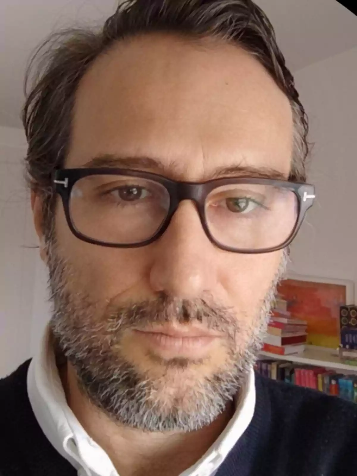 Hombre de cabello castaño y barba con gafas oscuras, vistiendo camisa blanca y suéter oscuro, con estantería de libros al fondo Hombre de cabello castaño y barba con gafas oscuras, vistiendo camisa blanca y suéter oscuro, con estantería de libros al fondo