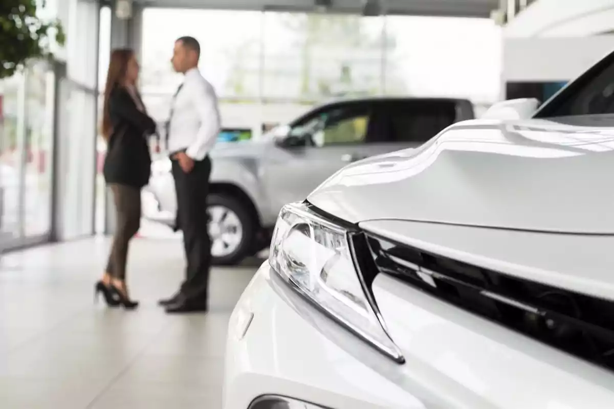 Frente de un auto blanco en primer plano con dos personas conversando en una agencia de autos al fondo Frente de un auto blanco en primer plano con dos personas conversando en una agencia de autos al fondo