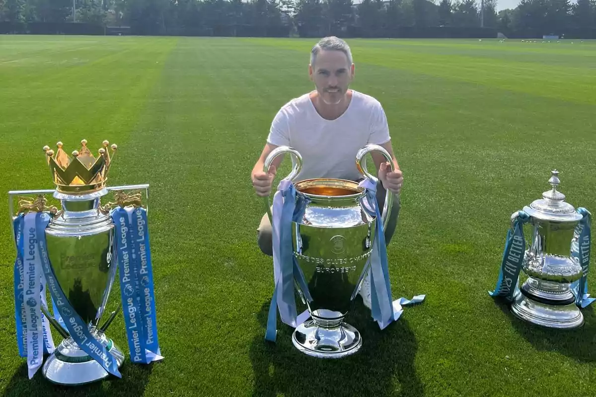 Hombre posando en un campo de fútbol con tres trofeos grandes de fútbol frente a él Hombre posando en un campo de fútbol con tres trofeos grandes de fútbol frente a él