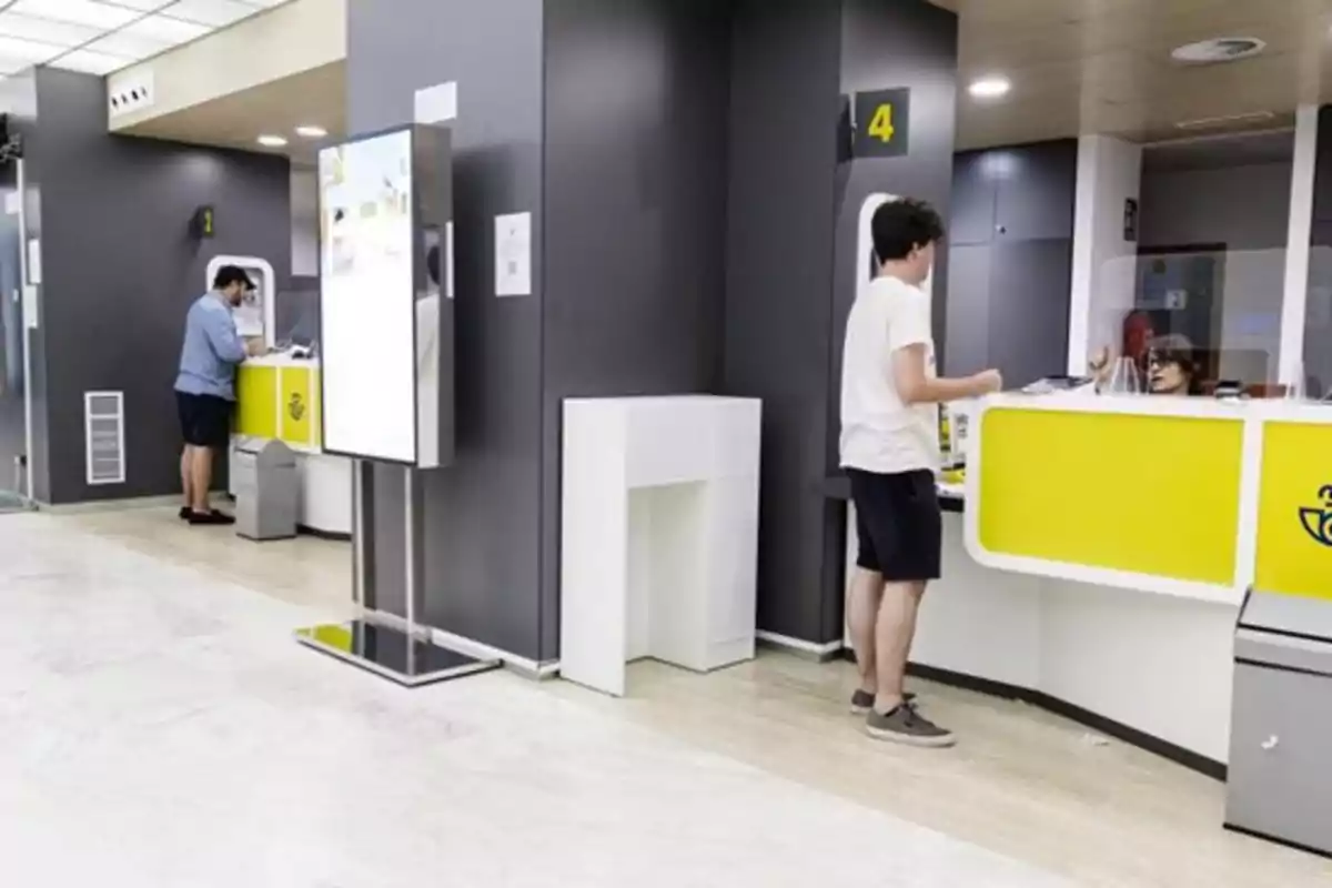 Oficina de correos moderna con dos personas siendo atendidas en mostradores amarillos y una empleada detrás de una mampara Oficina de correos moderna con dos personas siendo atendidas en mostradores amarillos y una empleada detrás de una mampara