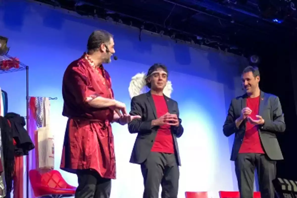 Tres hombres en un escenario, uno lleva bata roja y micrófono, otro tiene alas de ángel y aureola, el tercero sonríe, todos visten camisetas rojas y chaquetas oscuras