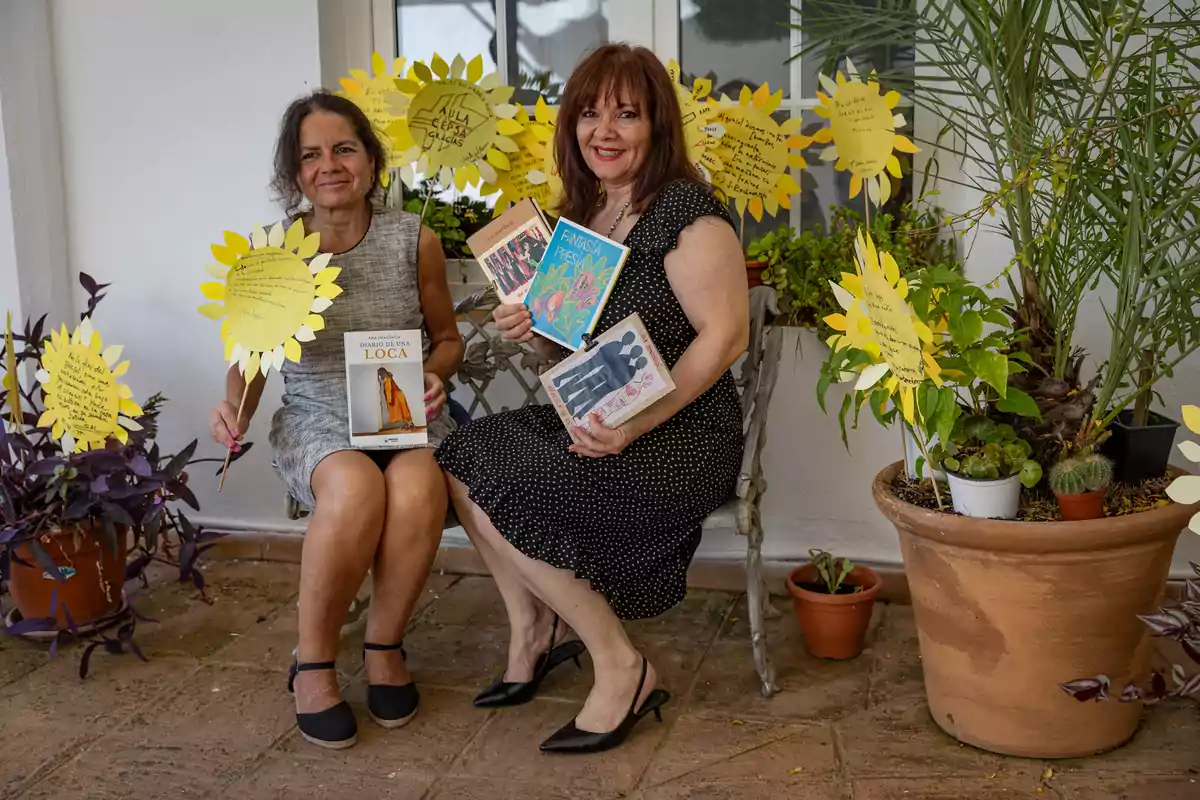 Dos mujeres sentadas en un banco rodeadas de plantas y girasoles de papel con mensajes escritos, cada una sostiene varios libros y sonríe a la cámara Dos mujeres sentadas en un banco rodeadas de plantas y girasoles de papel con mensajes escritos, cada una sostiene varios libros y sonríe a la cámara