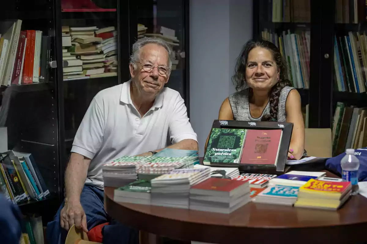 Dos personas sentadas en una mesa rodeada de libros en una biblioteca, con varias pilas de libros y revistas sobre la mesa Dos personas sentadas en una mesa rodeada de libros en una biblioteca, con varias pilas de libros y revistas sobre la mesa