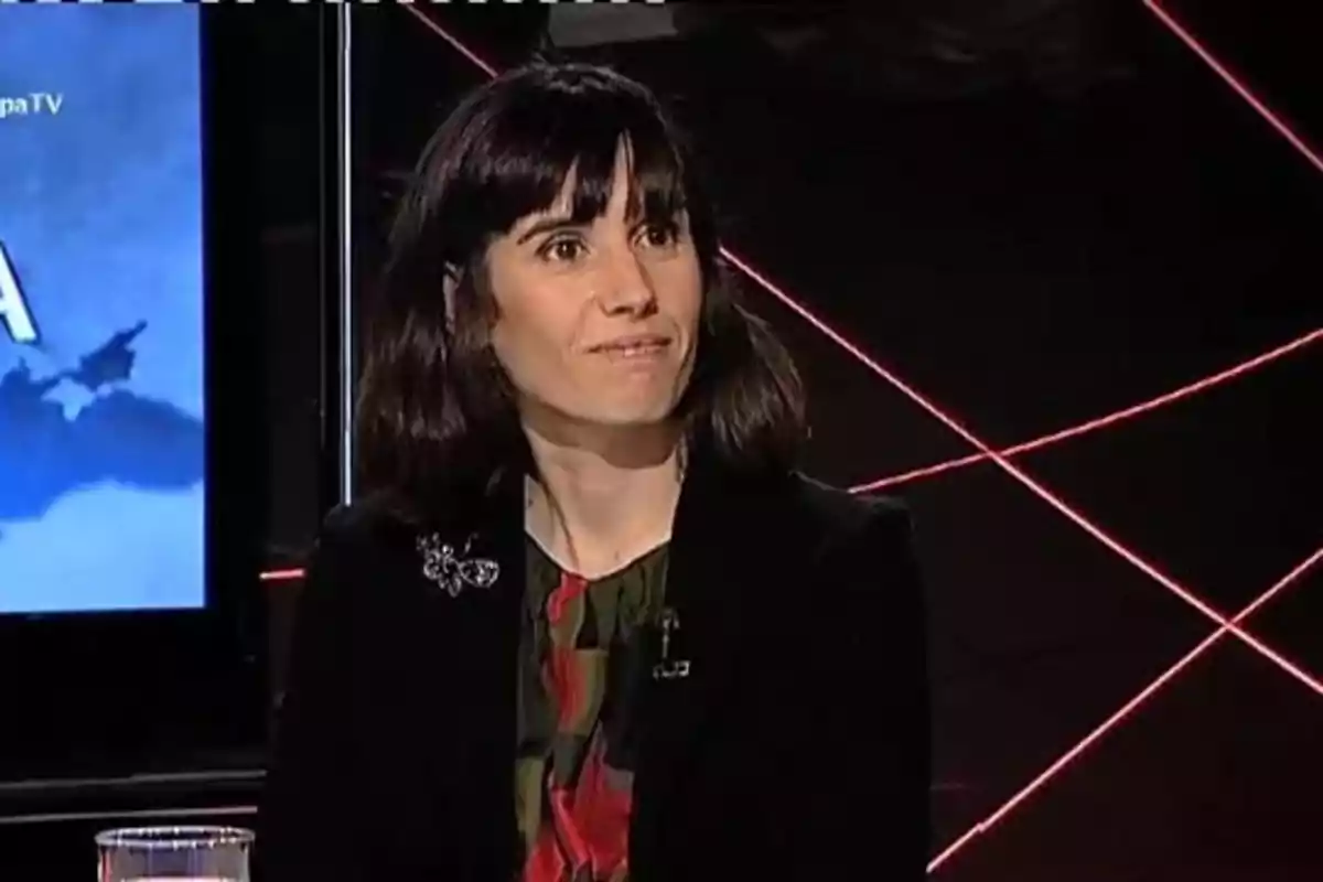 Mujer de cabello oscuro y liso sentada en un estudio de televisión con fondo oscuro y líneas rojas Mujer de cabello oscuro y liso sentada en un estudio de televisión con fondo oscuro y líneas rojas
