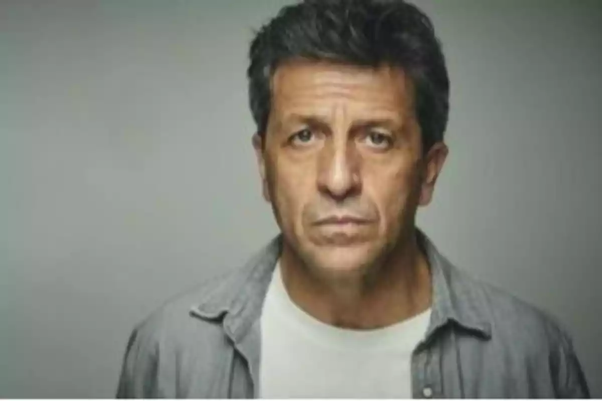 Hombre de cabello corto y oscuro con expresión seria vistiendo camiseta blanca y camisa gris frente a un fondo neutro Hombre de cabello corto y oscuro con expresión seria vistiendo camiseta blanca y camisa gris frente a un fondo neutro