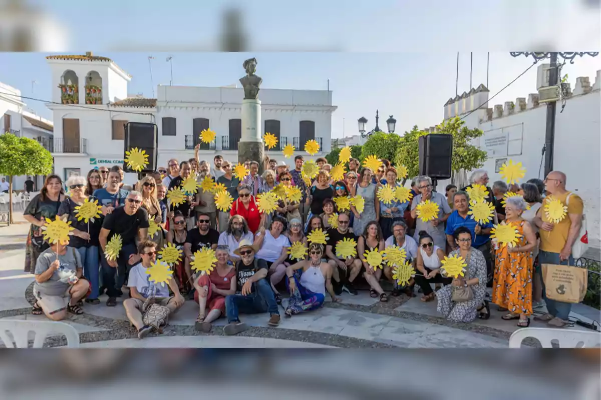 Un grupo numeroso de personas sonríe y sostiene carteles amarillos en forma de sol en una plaza soleada con edificios blancos al fondo
