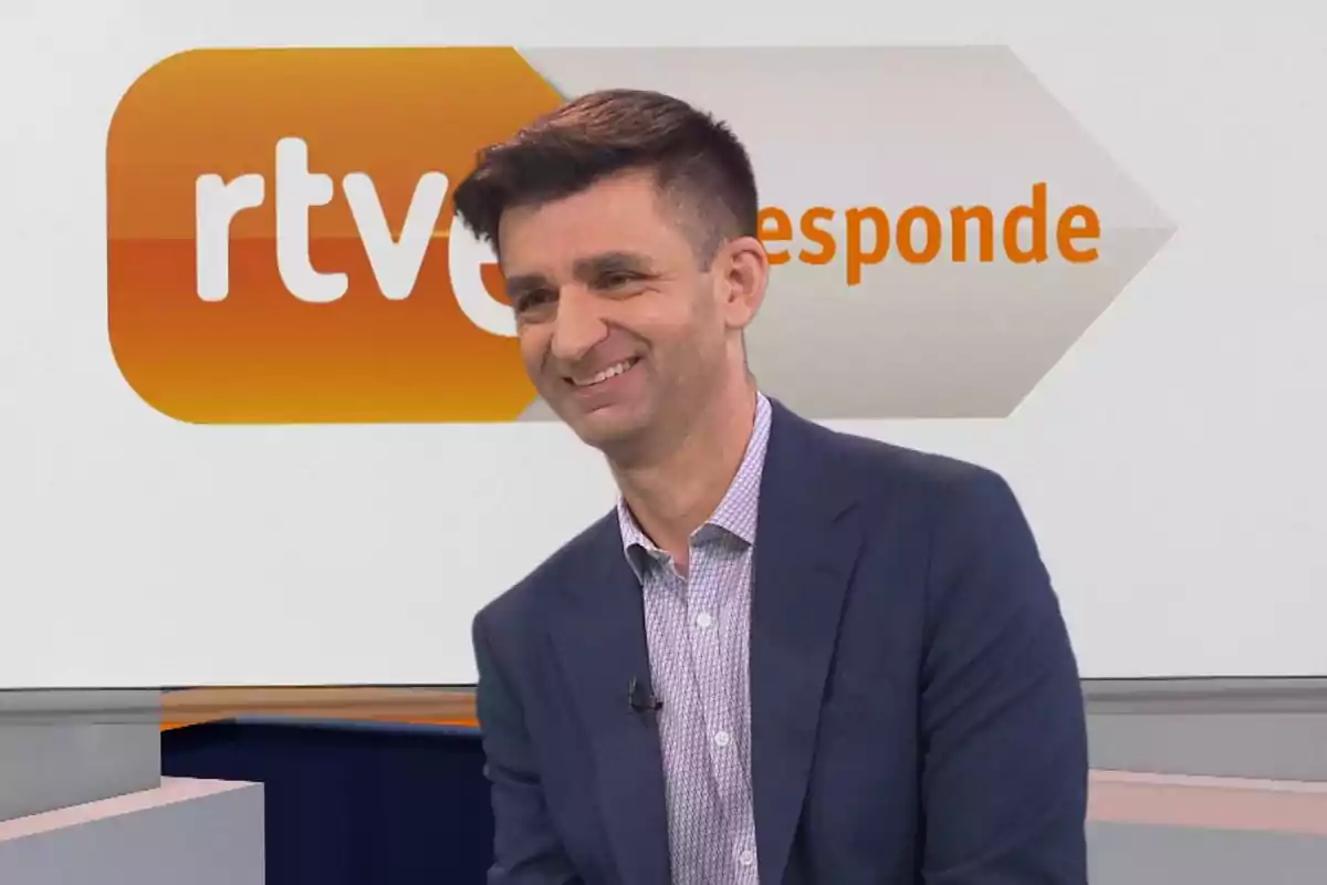Hombre con chaqueta azul y camisa clara sonriendo en un set de televisión con el logo de RTVE responde al fondo Hombre con chaqueta azul y camisa clara sonriendo en un set de televisión con el logo de RTVE responde al fondo