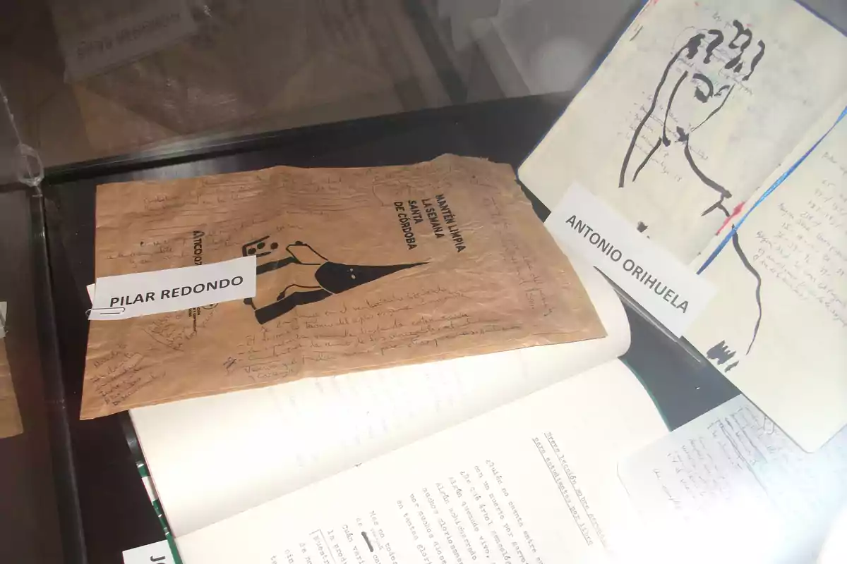 Exhibición de documentos y cuadernos manuscritos de Pilar Redondo y Antonio Orihuela en una vitrina, con etiquetas identificativas y textos escritos a mano y a máquina. Exhibición de documentos y cuadernos manuscritos de Pilar Redondo y Antonio Orihuela en una vitrina, con etiquetas identificativas y textos escritos a mano y a máquina.