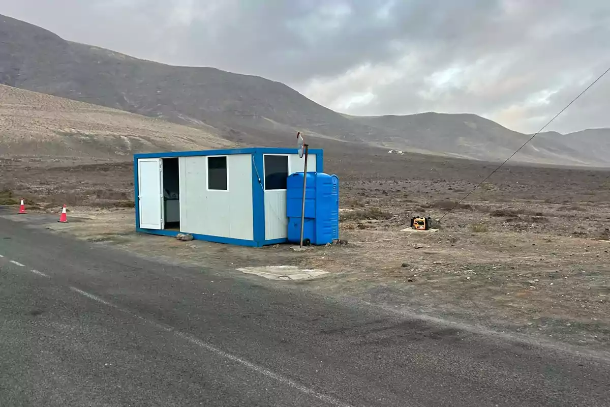 Caseta blanca con bordes azules y baño portátil azul junto a una carretera en un paisaje árido y montañoso Caseta blanca con bordes azules y baño portátil azul junto a una carretera en un paisaje árido y montañoso
