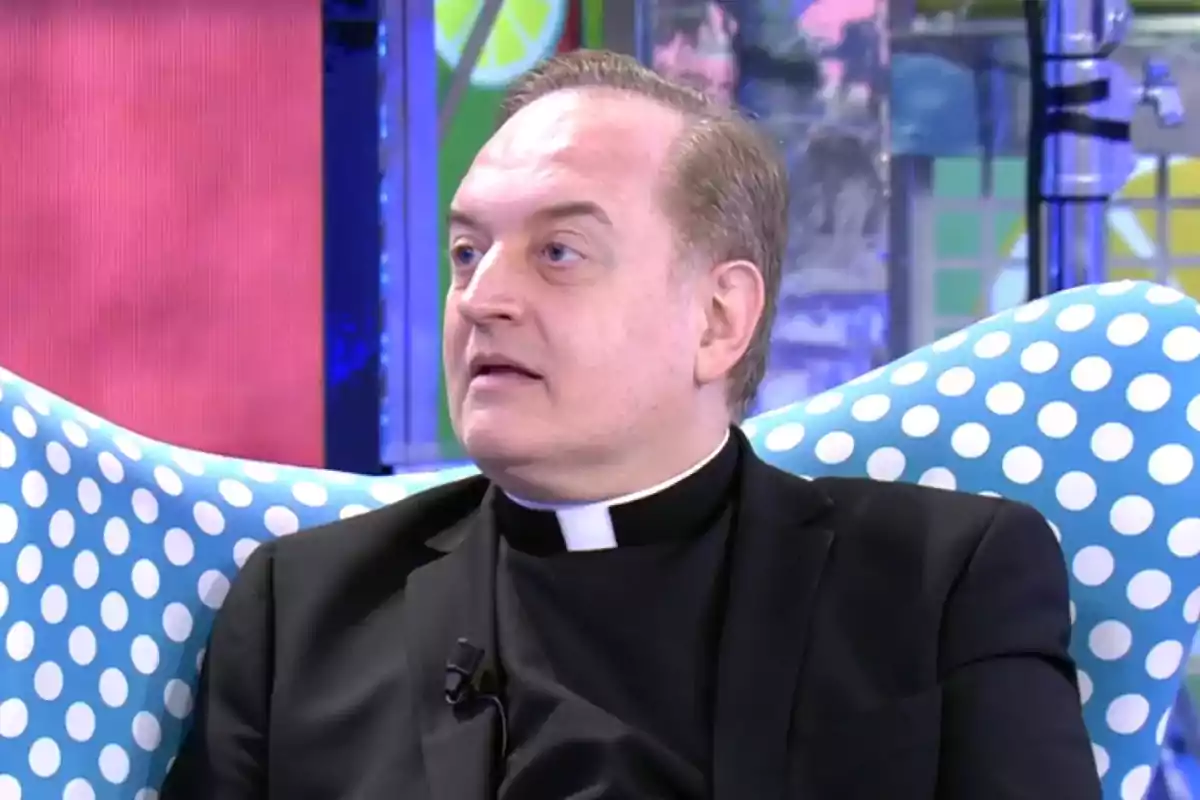 Hombre con sotana negra y alzacuellos blanco sentado en un sillón azul con lunares blancos en un set de televisión colorido Hombre con sotana negra y alzacuellos blanco sentado en un sillón azul con lunares blancos en un set de televisión colorido
