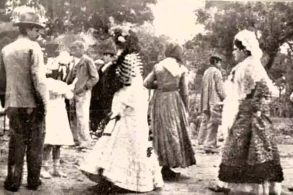 Personas vestidas con trajes tradicionales bailando y conversando al aire libre en una fotografía antigua en blanco y negro Personas vestidas con trajes tradicionales bailando y conversando al aire libre en una fotografía antigua en blanco y negro