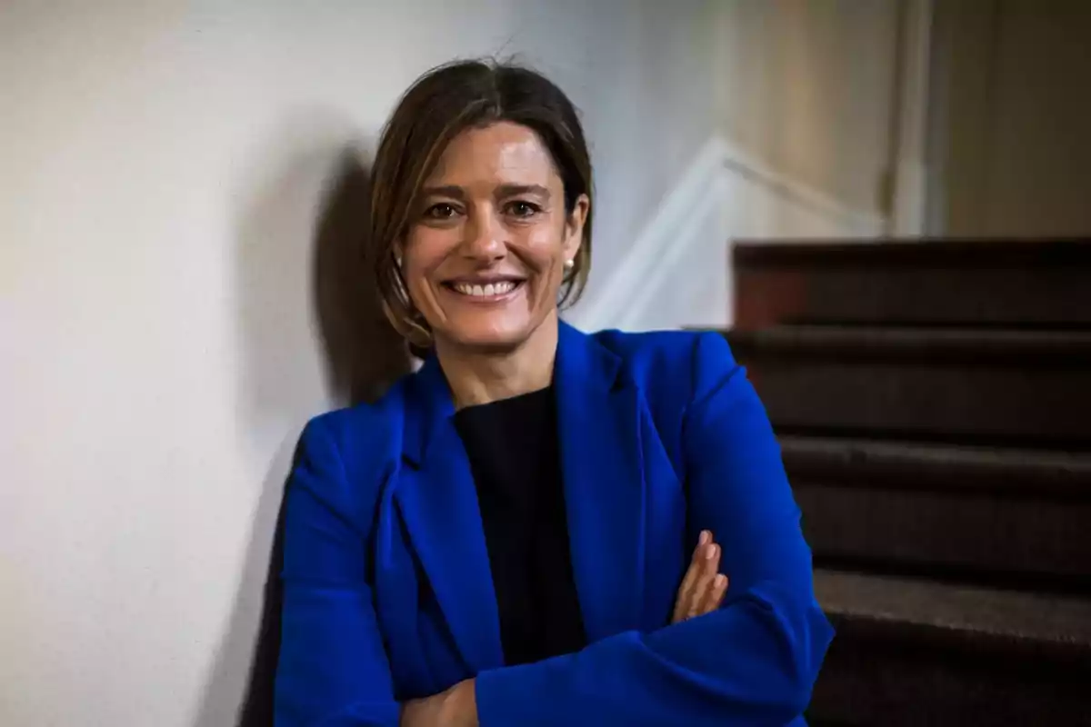 Mujer sonriendo con los brazos cruzados vestida con saco azul junto a una escalera en interiores Mujer sonriendo con los brazos cruzados vestida con saco azul junto a una escalera en interiores
